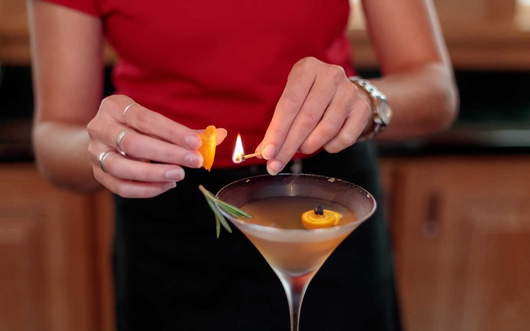 Woman squeezing an orange wedge to a flame of a match on top of a drink served in a martini glass with rosemary garnish and an orange wedge.