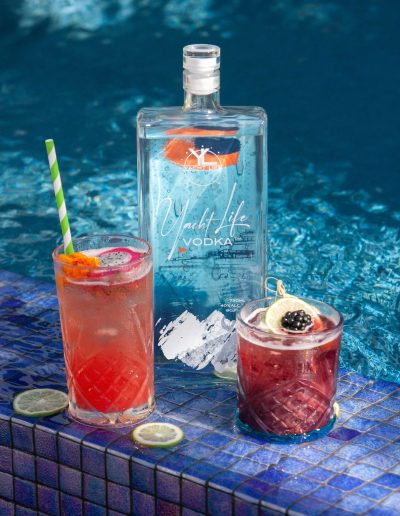 Yacht Life Vodka bottle on edge of a pool with two drinks in front of it.
