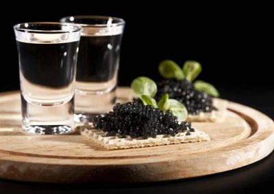 Caviar on a waffle cracker served on a wooden tray with two shot glasses of vodka.