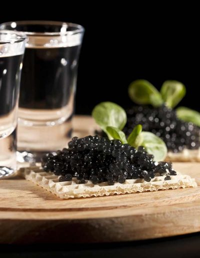 Caviar on a waffle cracker served on a wooden tray with two shot glasses of vodka.