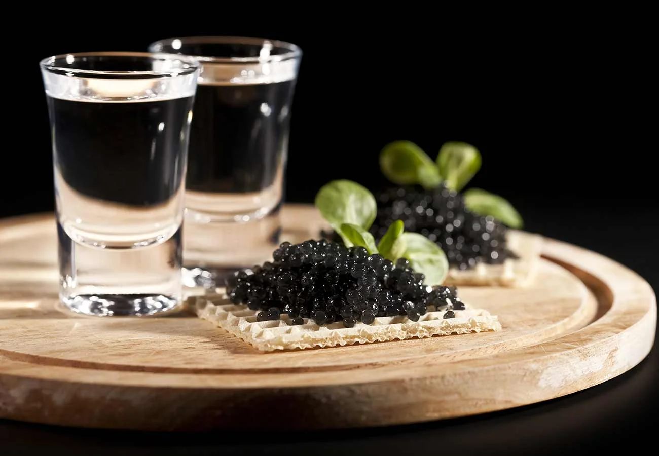 Caviar on a waffle cracker served on a wooden tray with two shot glasses of vodka.