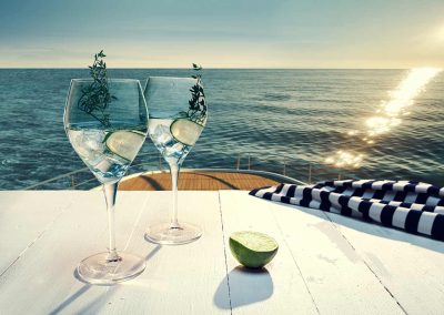 Clear beverage served in a wine glass, on top of a table that is on top level of a yacht, looking down onto the sea and horizon.