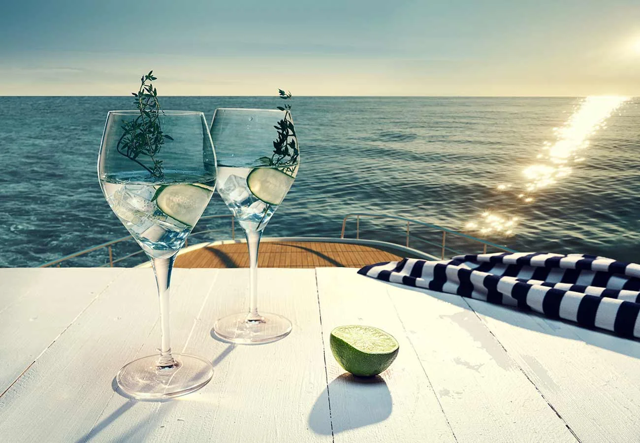 Clear beverage served in a wine glass, on top of a table that is on top level of a yacht, looking down onto the sea and horizon.