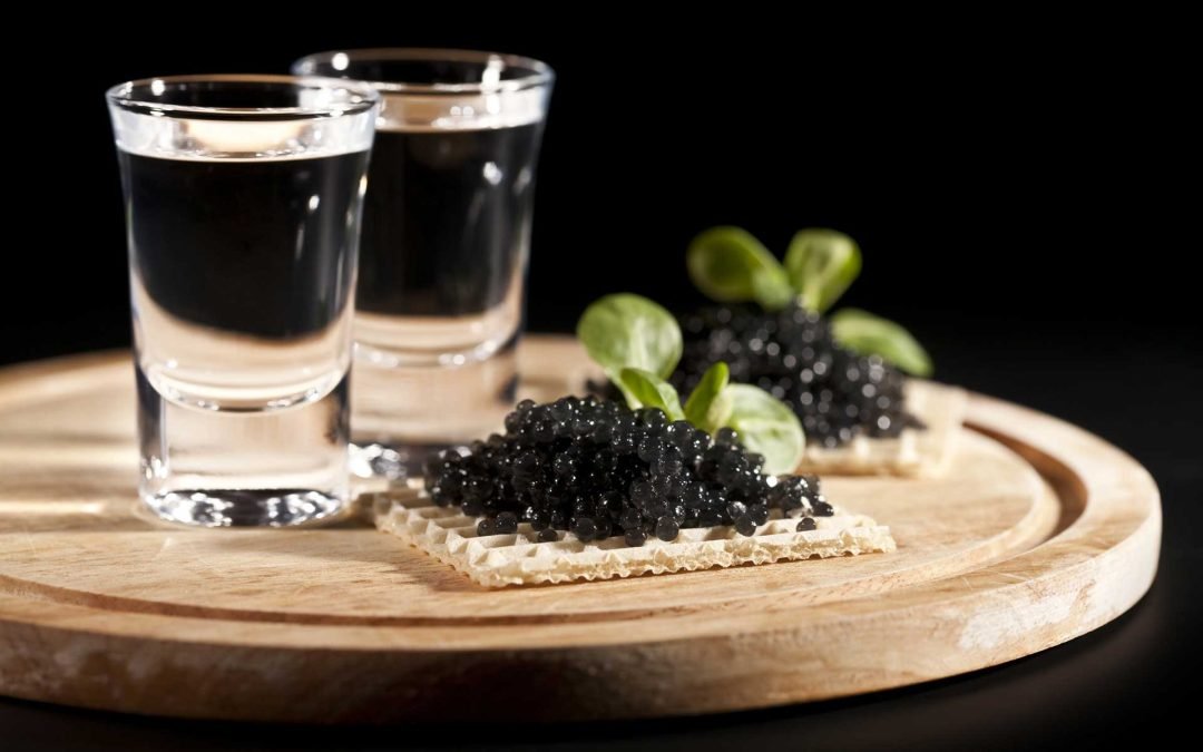 Vodka in two shot glasses behind a cracker with black caviar on it, displayed on a wooden board