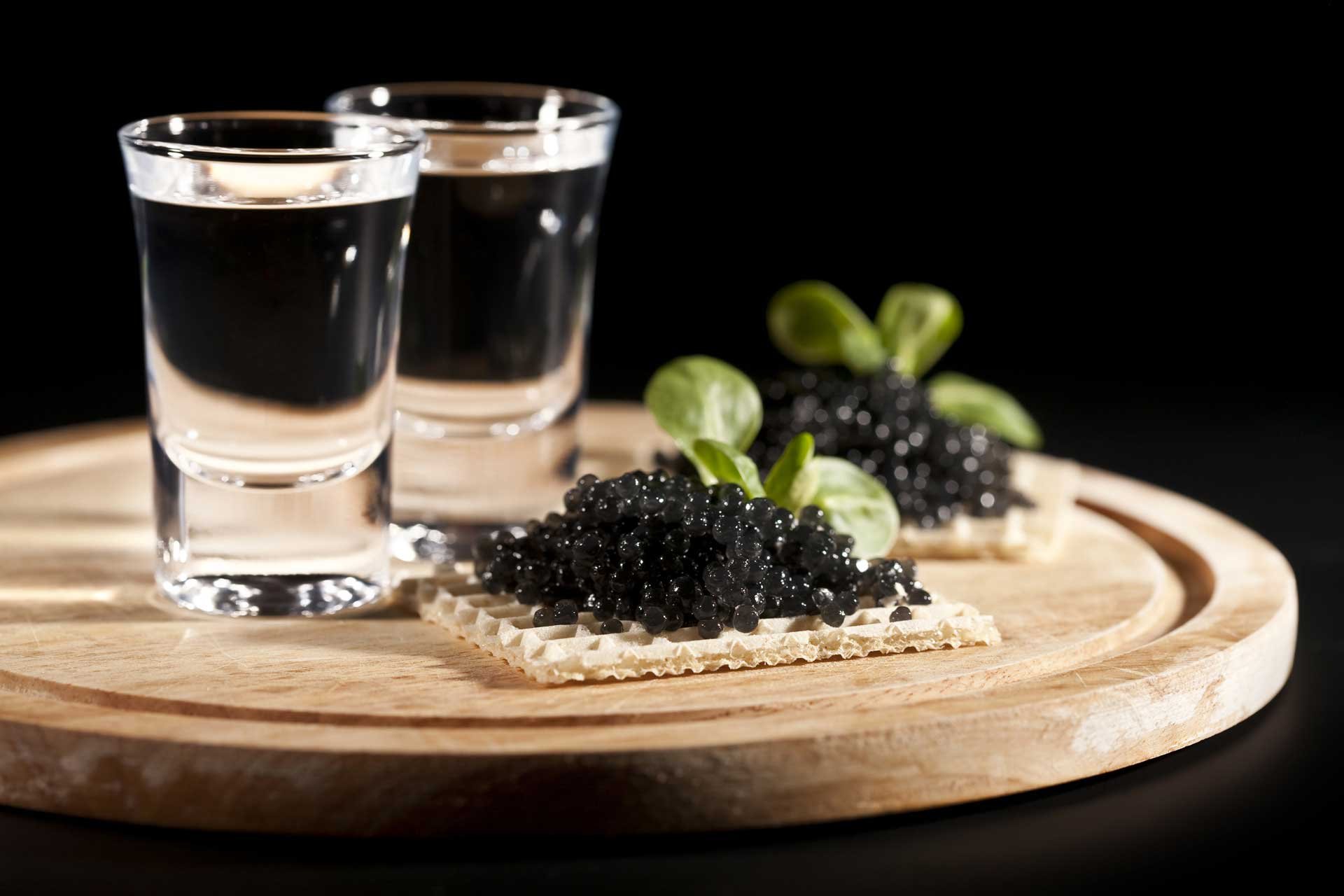 Vodka in two shot glasses behind a cracker with black caviar on it, displayed on a wooden board
